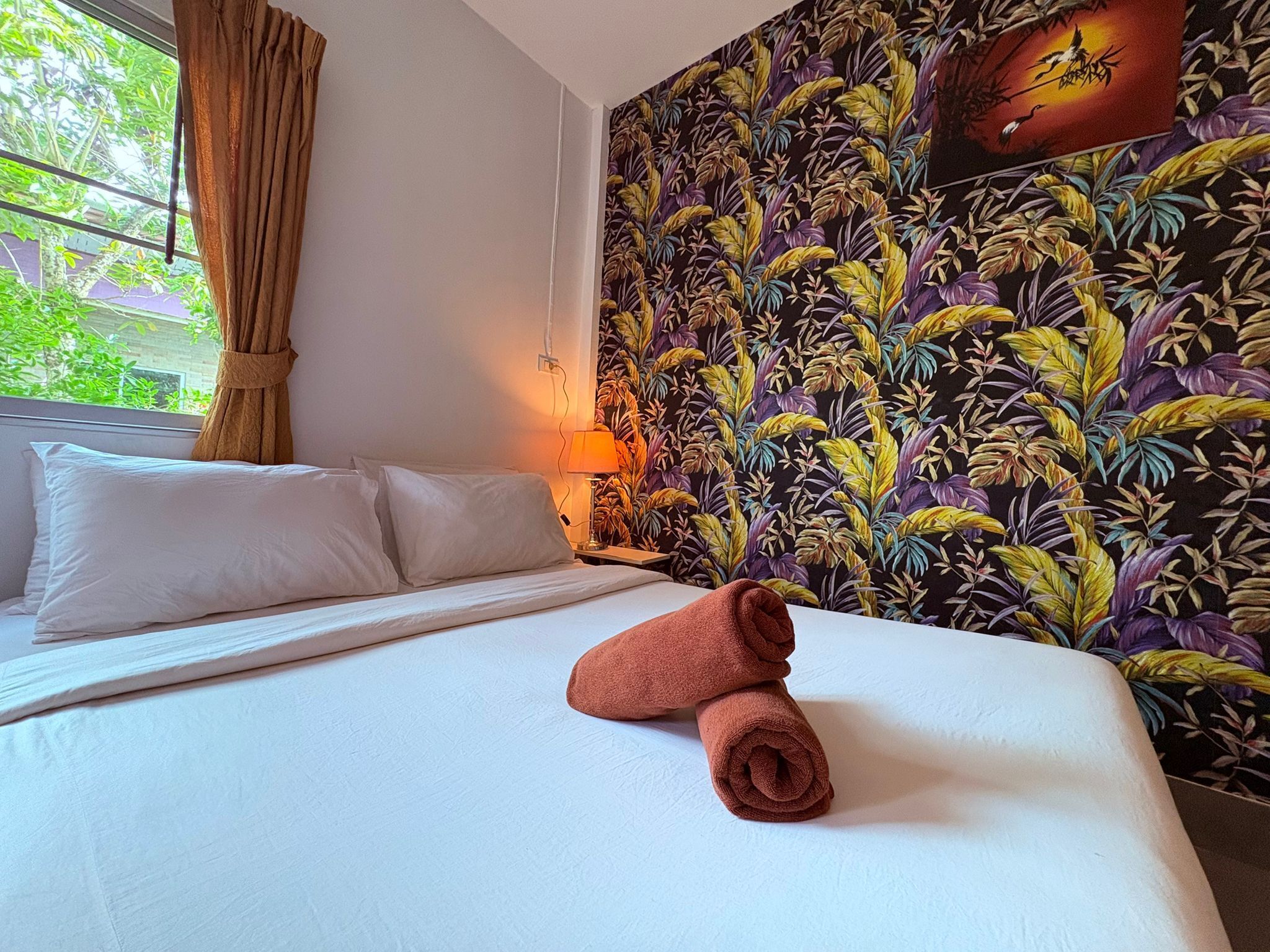 A hotel room with a bed, pillows, a rolled towel, a window with curtains, a wall with a tropical leaf pattern, and a wall-mounted lamp.