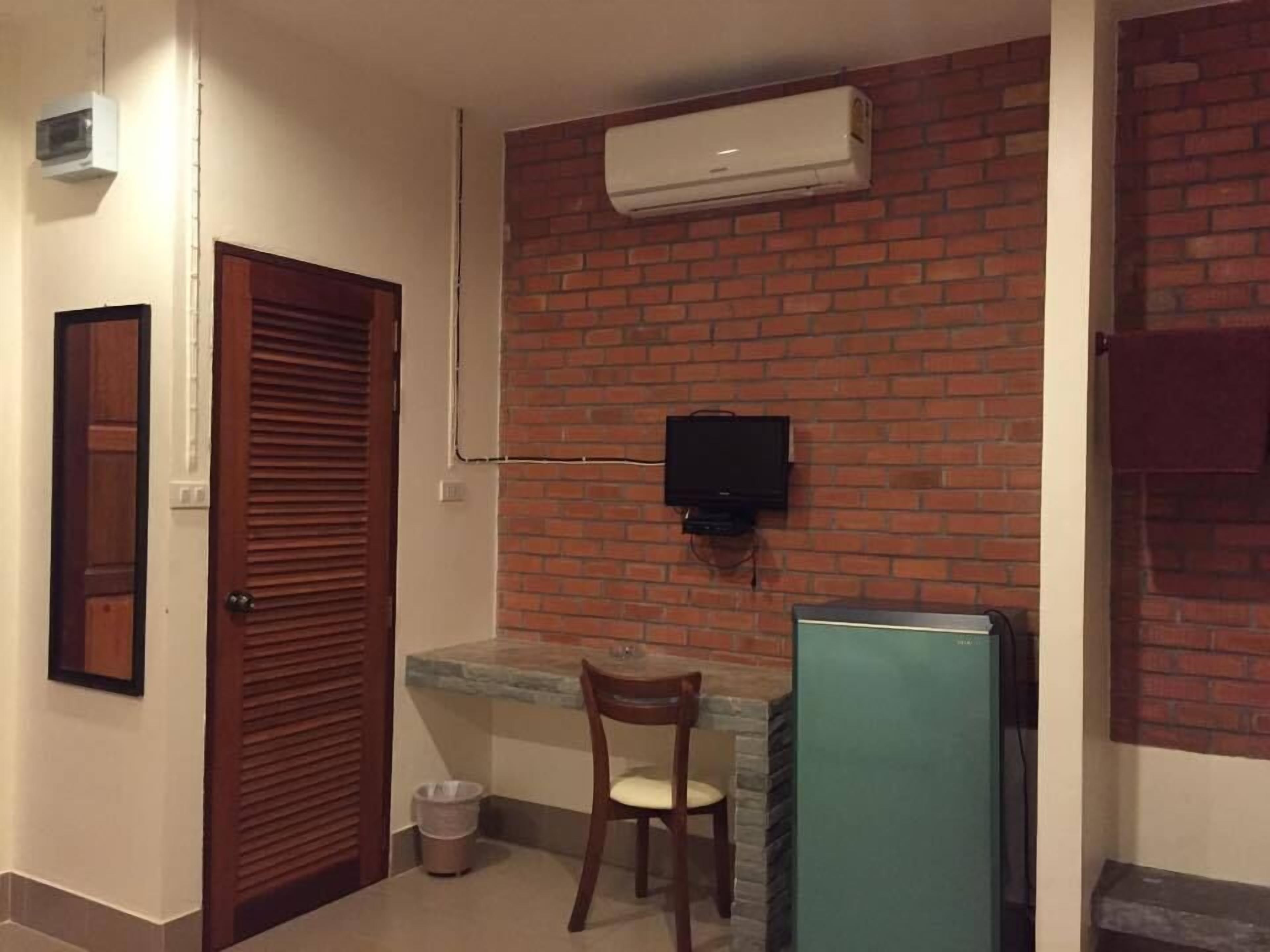 A small room with a brick wall, a wooden door, a small table with a chair, a TV mounted on the wall, and a refrigerator.