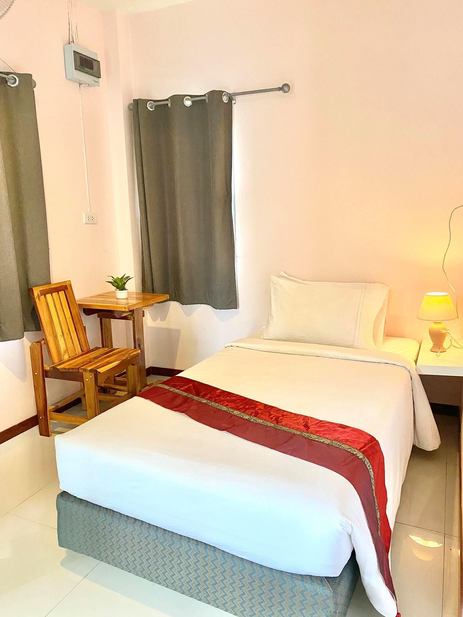 Ein Hotelzimmer mit einem Bett, einem Holzsessel, einem kleinen Tisch mit Pflanze, einem Nachttisch mit Lampe und einem an der Wand montierten Thermostat.
