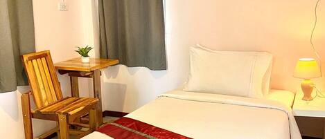 Ein Hotelzimmer mit einem Bett, einem Holzsessel, einem kleinen Tisch mit Pflanze, einem Nachttisch mit Lampe und einem an der Wand montierten Thermostat.
