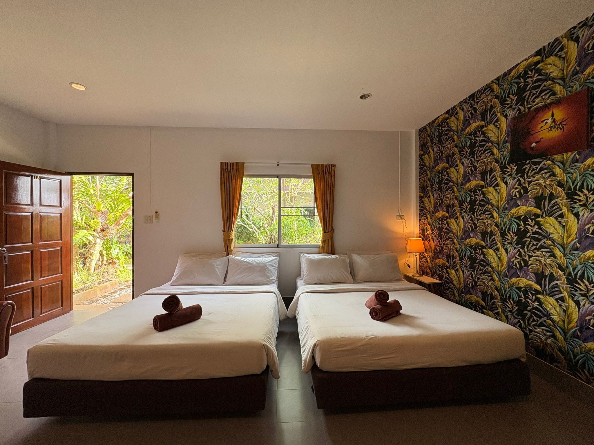 A room with two beds, a window with curtains, a door, and a wall with a floral pattern.