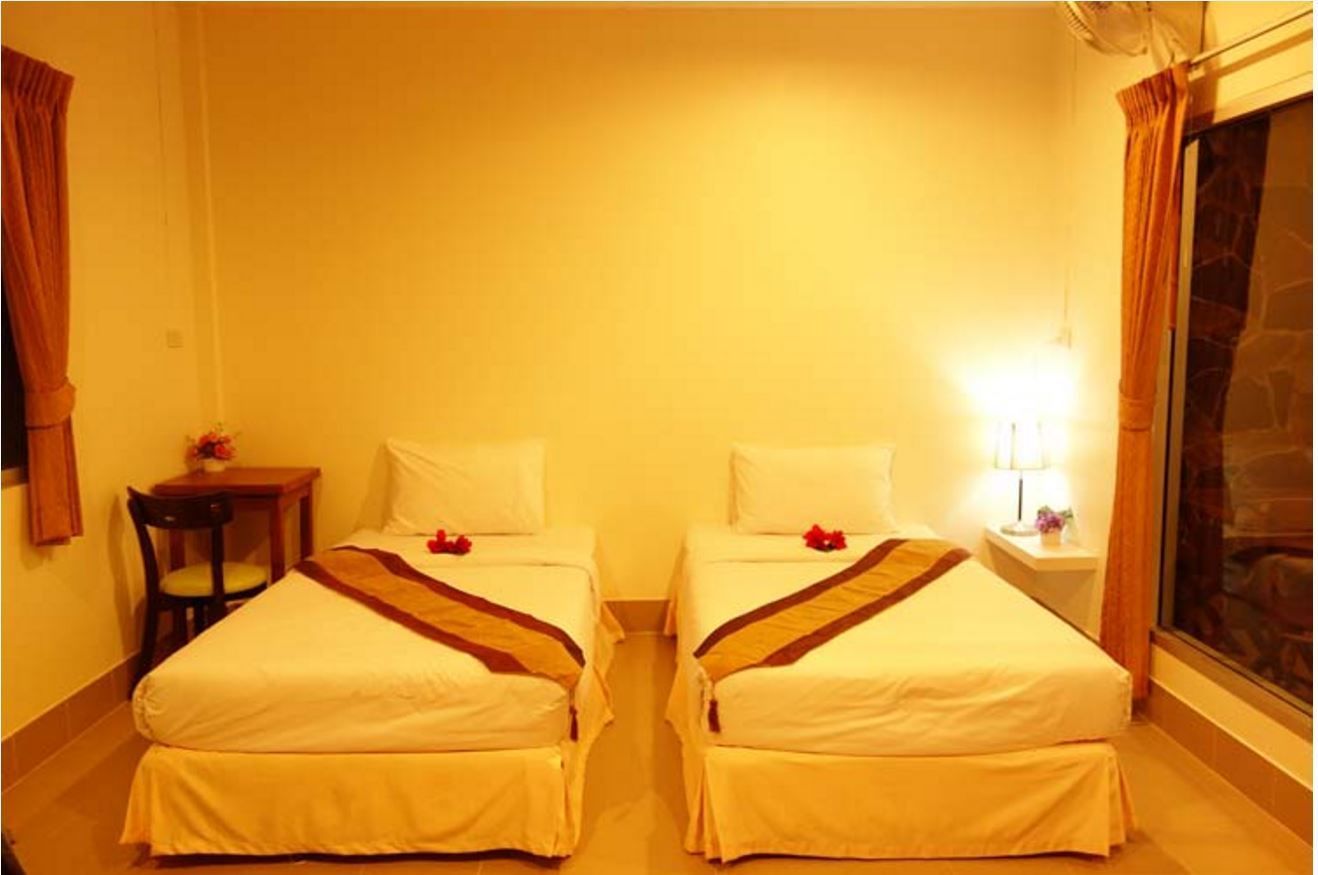 A hotel room with two single beds, a small table with a chair, a nightstand with a lamp, and a window with curtains.