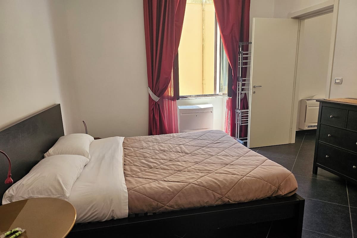 Een bed met een sprei, een nachtkastje, een kledingkast, een radiator en een raam met gordijnen.