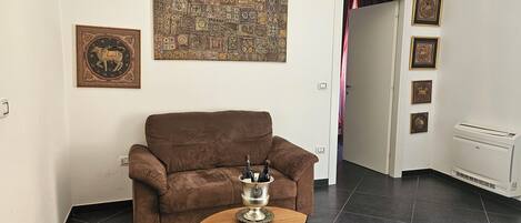 A living room with a brown sofa, a small wooden table with a silver container, a large patterned artwork, and a radiator.