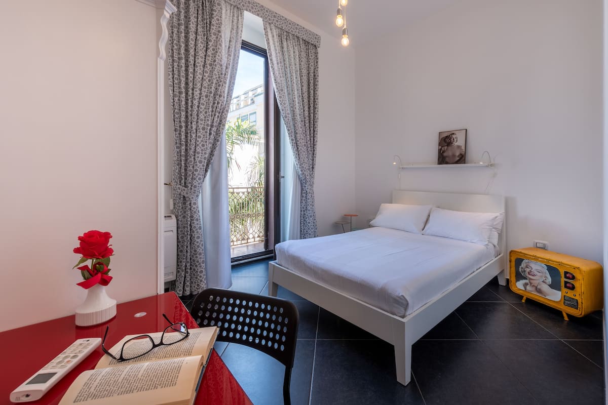 Een slaapkamer met een bed, een bureau met een computer, een stoel, een vaas met een rode roos en een klein tafeltje met een afstandsbediening en een boek.
