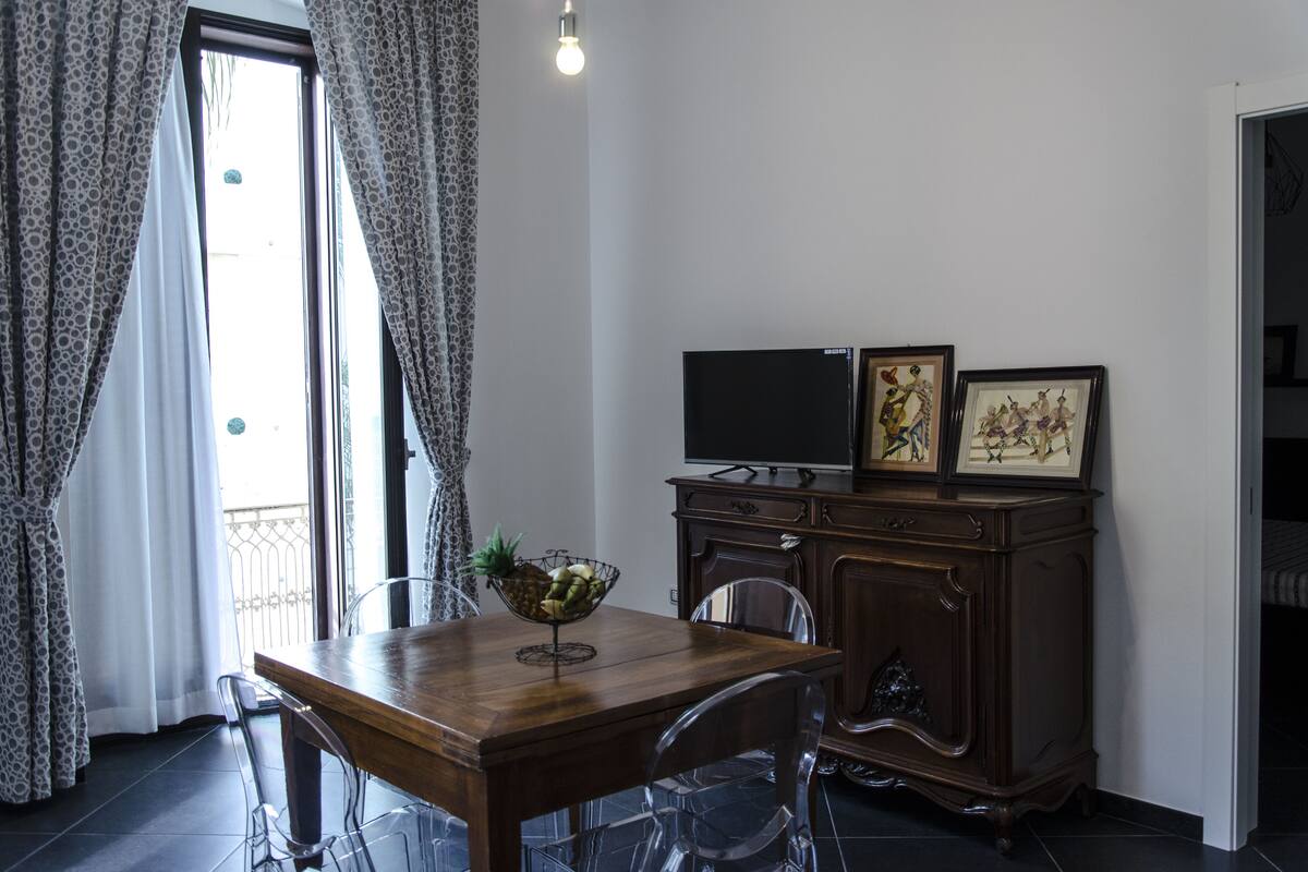 Een zithoek met een houten tafel, transparante stoelen, een schaal fruit, een donker houten kast met een flatscreen televisie en ingekaderde foto's, en een balkon met decoratieve gordijnen.