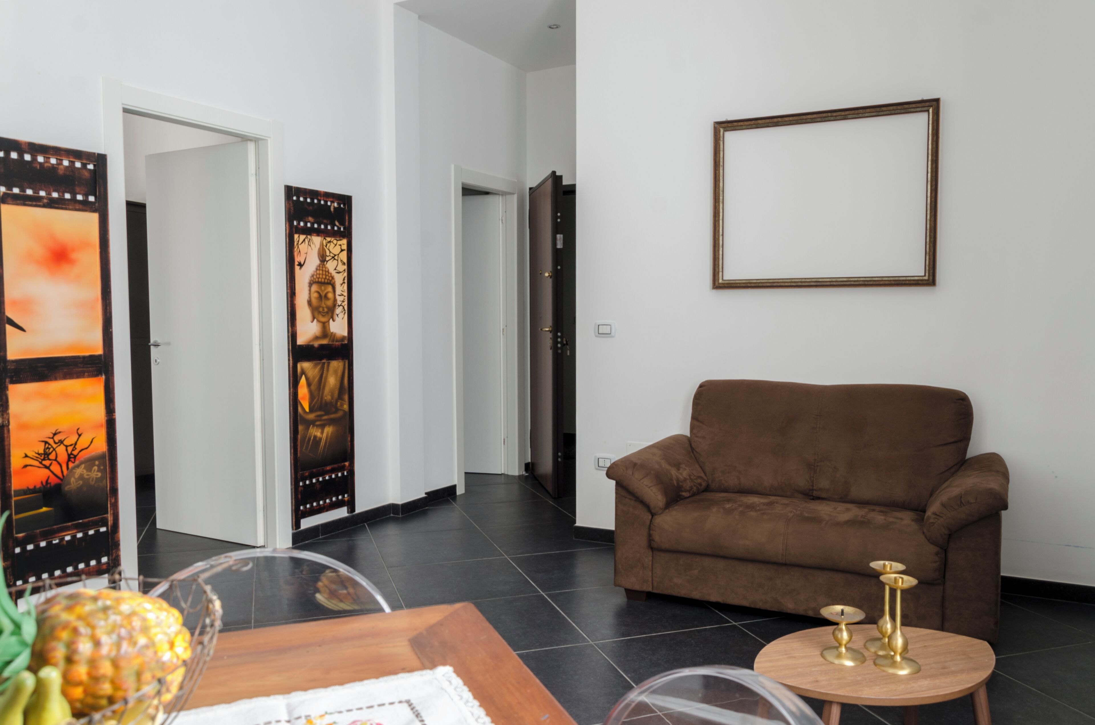 Een woonkamer met een bruine bank, een klein houten tafeltje met decoratieve items, en een aan de muur bevestigd fotolijstje.