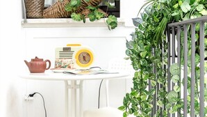 A small white table with a radio, a teapot, and a cup. There is a chair in front of the table, and a window with a plant on the sill.
