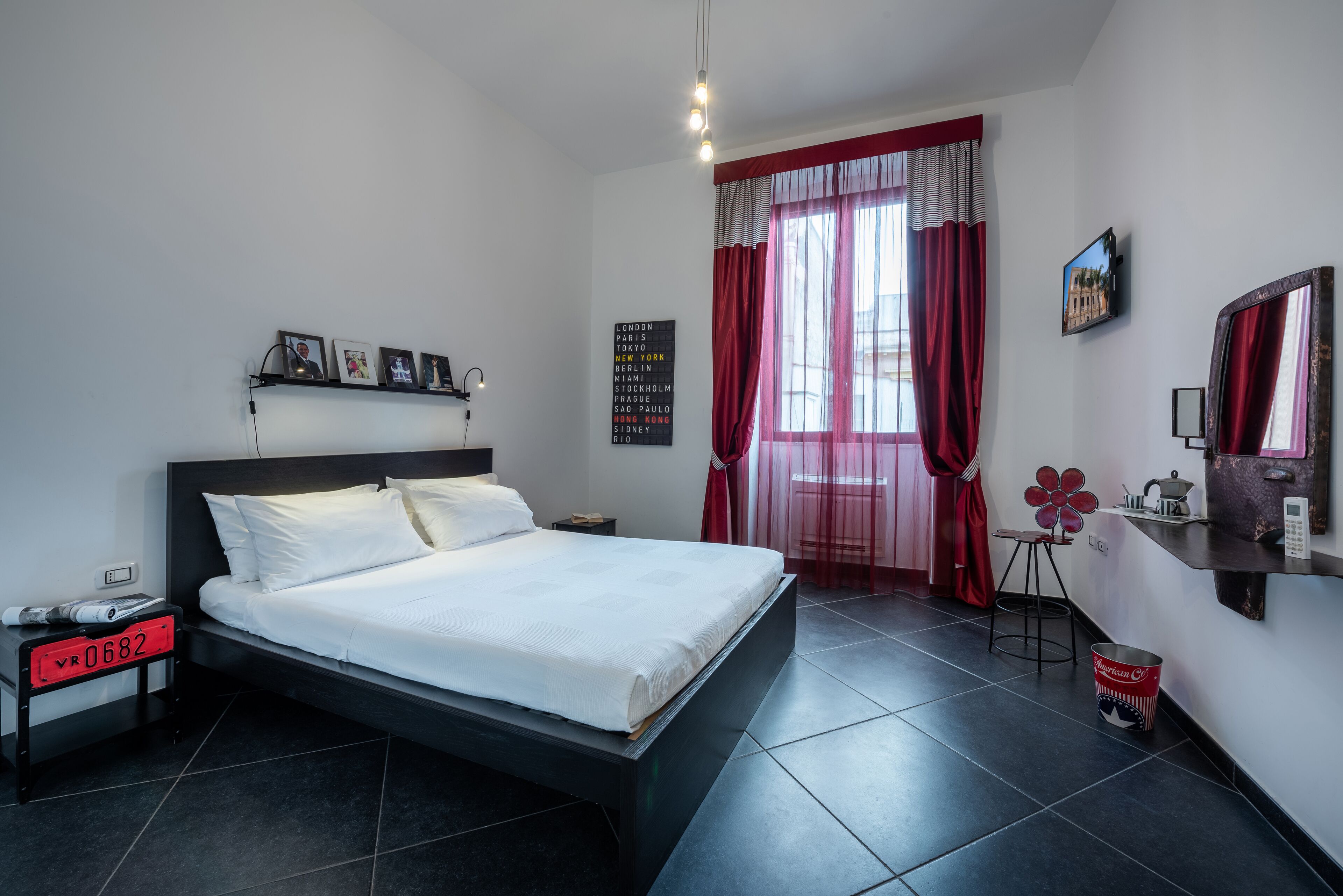 A modern bedroom with a large bed, a nightstand, a desk with a chair, and a window with red curtains.