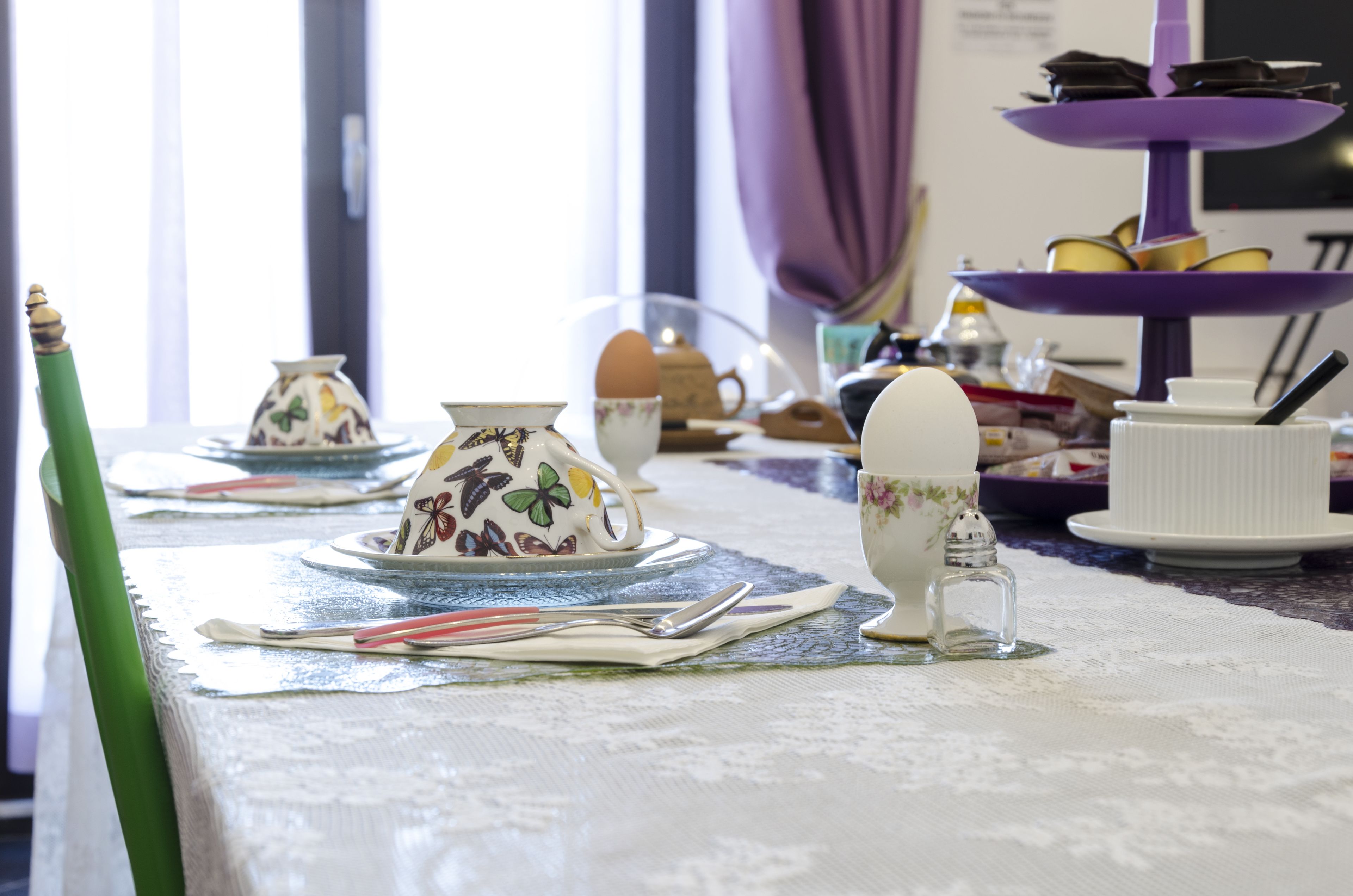 Uma mesa posta com xícaras de chá, pires e um bule, com um suporte roxo em vários níveis ao fundo.