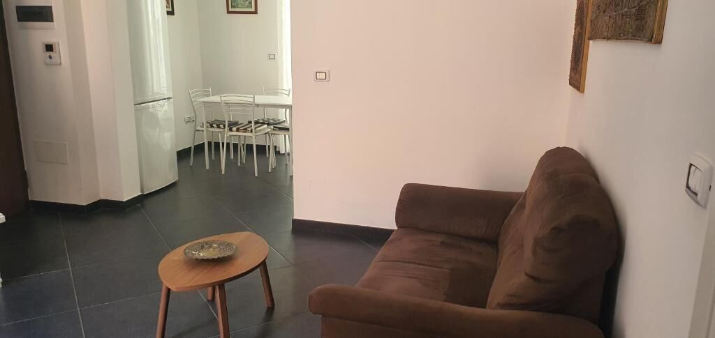 A living room with a brown sofa, a small wooden table, and a dining area with white chairs and a table.