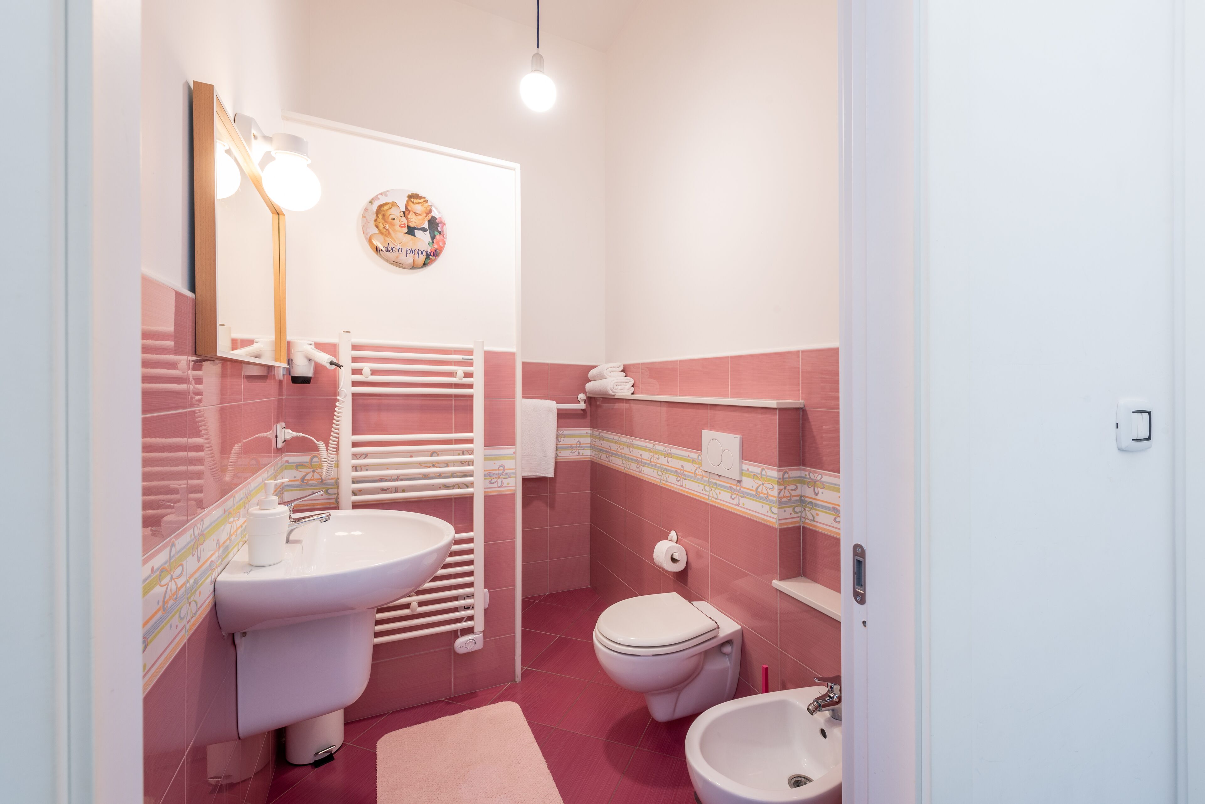 A bathroom with a white sink, toilet, and bidet, a towel rack, and a mirror with lights.