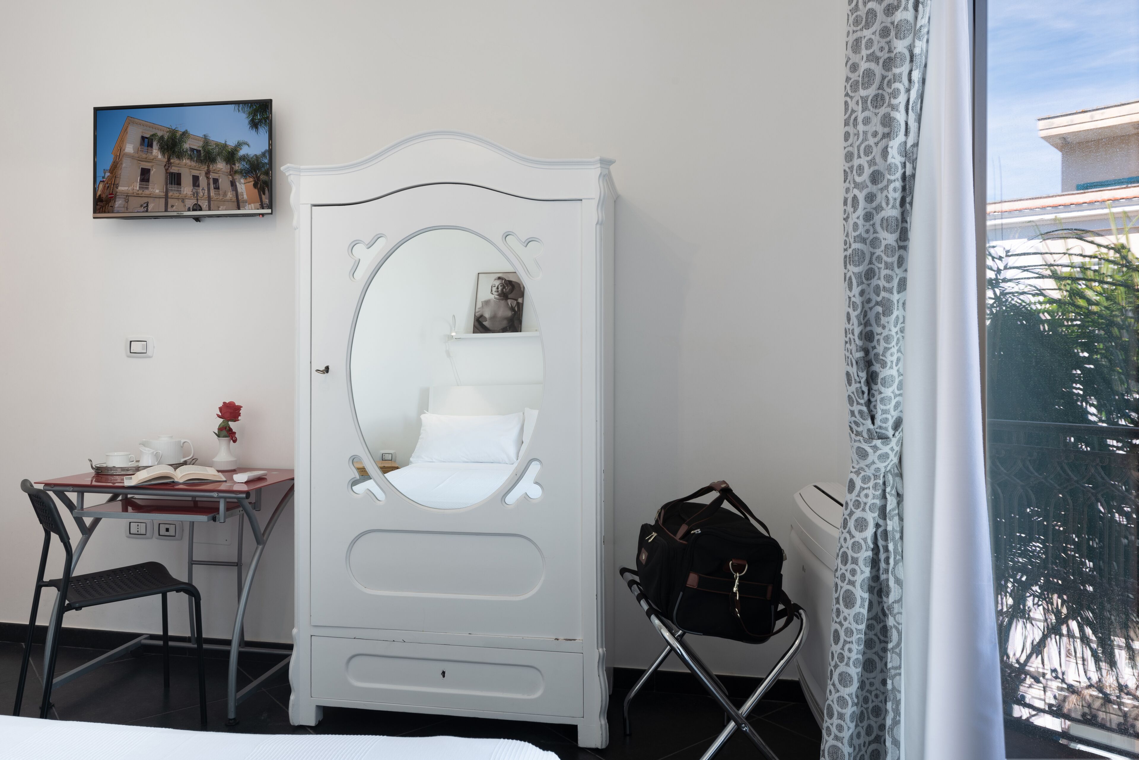 A bedroom with a white wardrobe, a desk with a chair, a bed, a window with a view of palm trees, and a television on the wall.