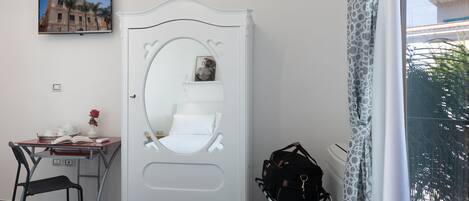 A bedroom with a white wardrobe, a desk with a chair, a bed, a window with a view of palm trees, and a television on the wall.