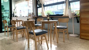 A cafe with wooden chairs and tables, large windows with curtains, and a view of the street outside. - Sleep Sheep Phuket Hostel SHA (Phuket)