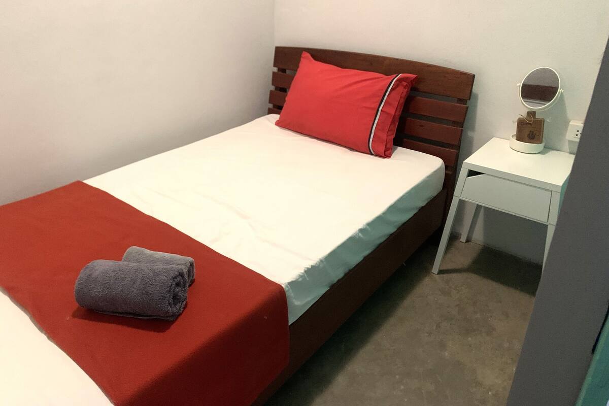 A single bed with a red blanket and a pillow, a small bedside table with a lamp, and a wall-mounted mirror.