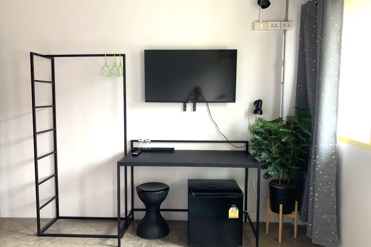 A compact workspace with a desk, a small trash bin, a ladder, a potted plant, and a TV mounted on the wall.