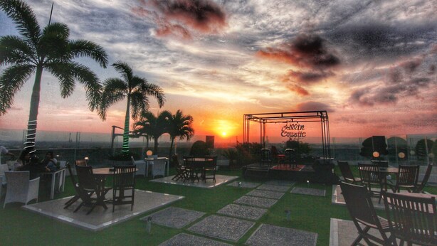 An outdoor seating area with tables and chairs, palm trees, and a stage with a sign.
