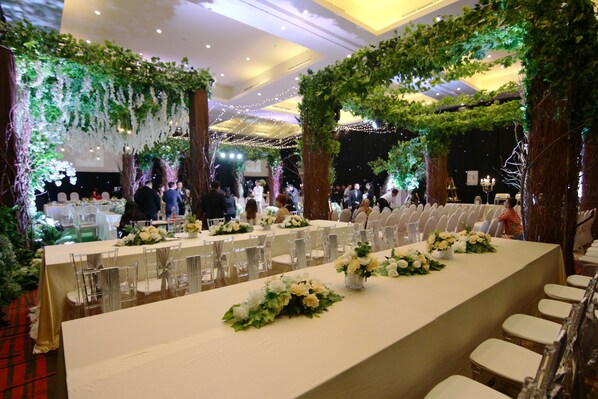 A banquet hall with long tables, clear chairs, and greenery decorations.