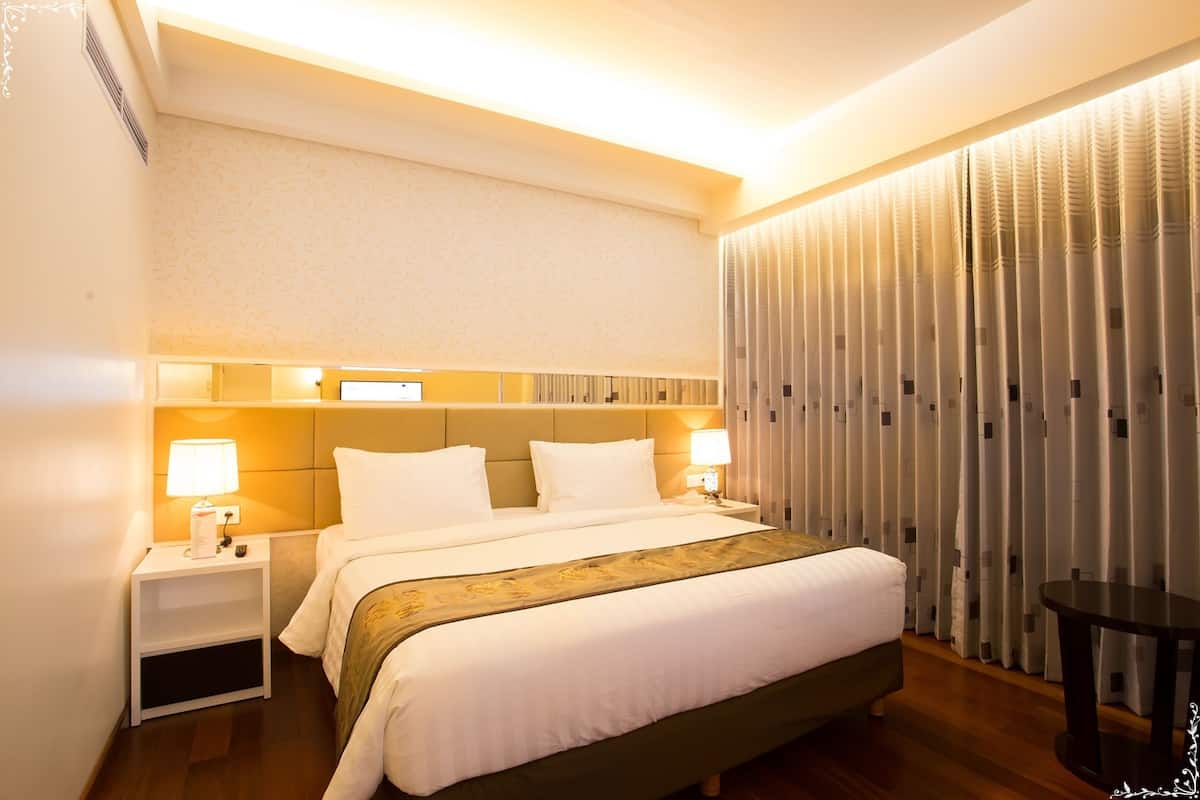 Una habitación de hotel con una cama grande, dos mesitas de noche con lámparas, una pequeña mesa y un ventanal cubierto por una cortina.