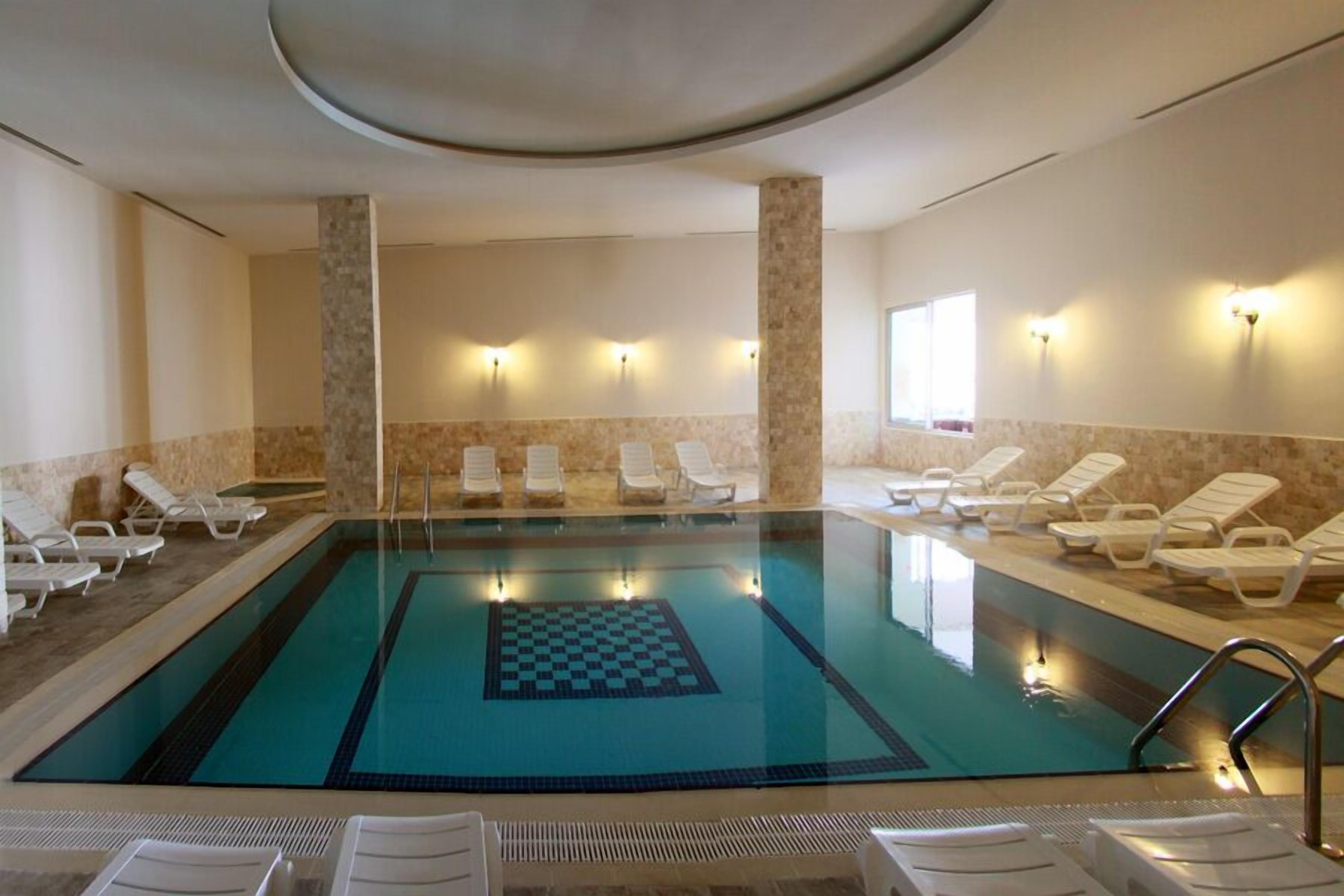 A swimming pool with a checkered pattern in the center, surrounded by lounge chairs and walls with light fixtures.
