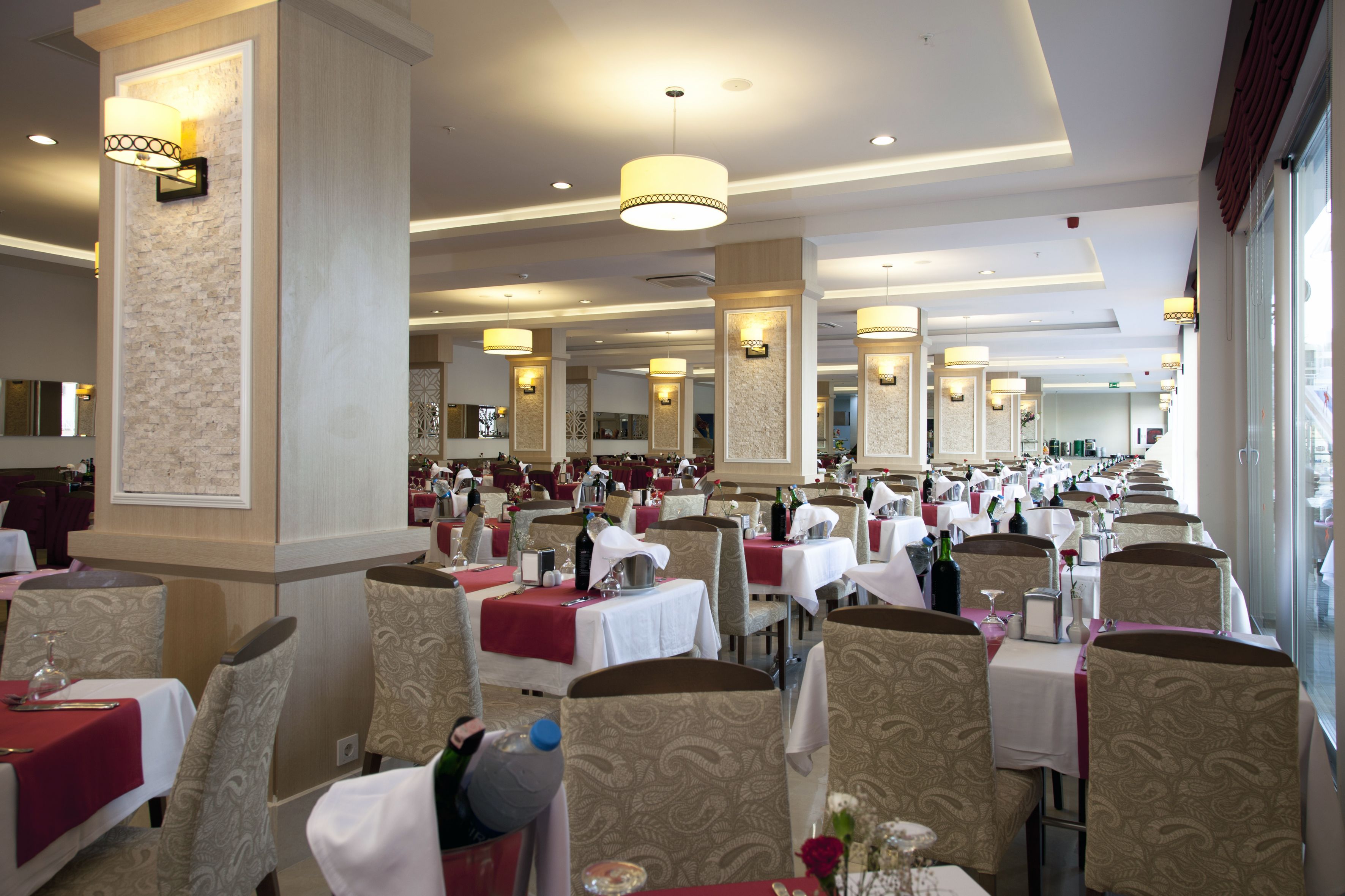 A spacious dining area with multiple tables and chairs, well-lit by ceiling lights, and featuring a modern interior design with light-colored walls and patterned upholstery.