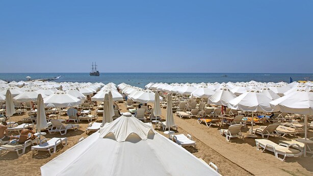 A beach with rows of white umbrellas and sun loungers, a clear blue sky, and a ship in the distance. - Merve Sun Hotel & Spa - All Inclusive (Manavgat)