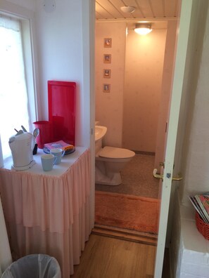 A bathroom with a toilet, a sink, and a shower area. There is a window with curtains, a trash bin, and a small table with a red container and a white pitcher.