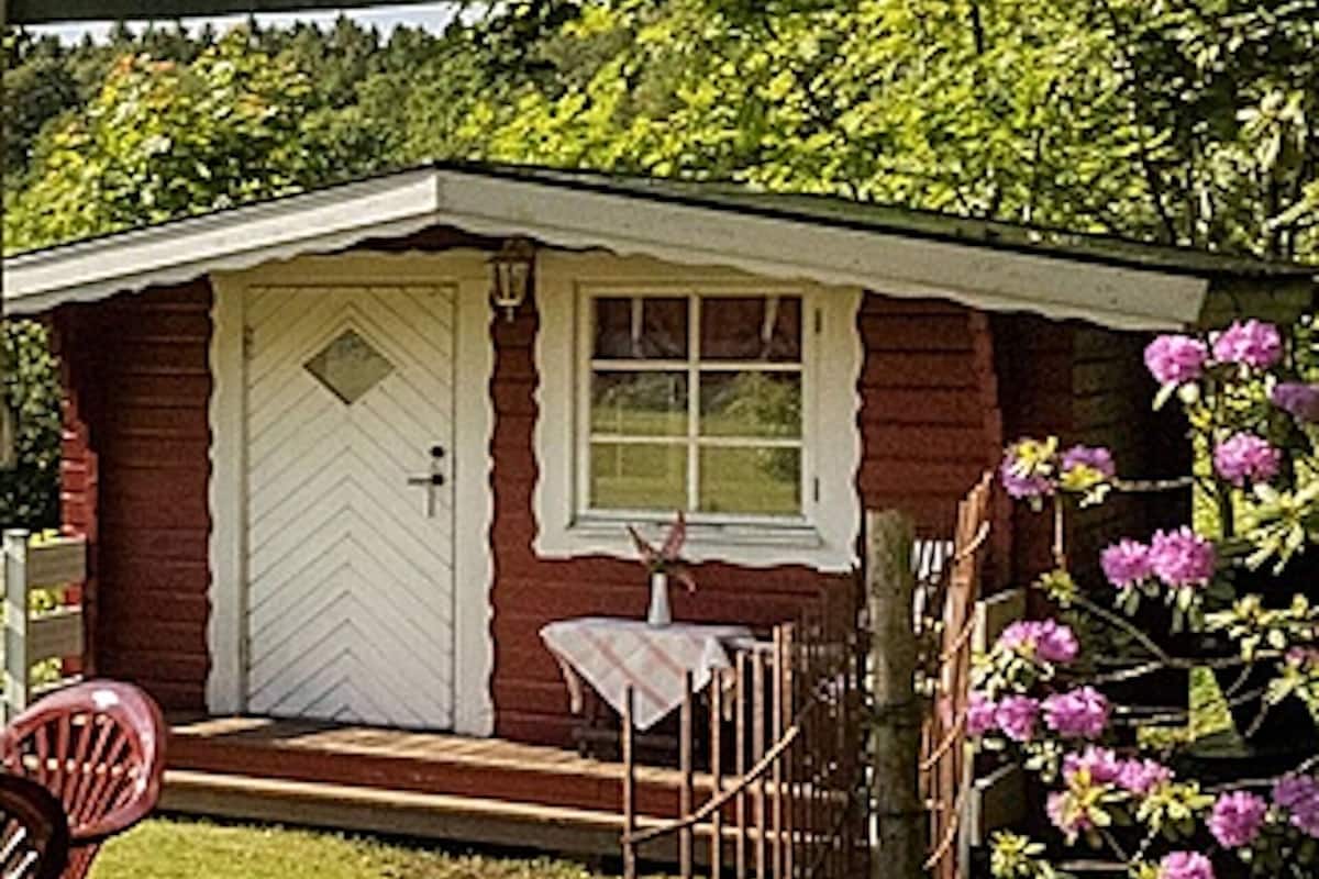 A small red house with a white door and window, a small porch with a table, and a chair.