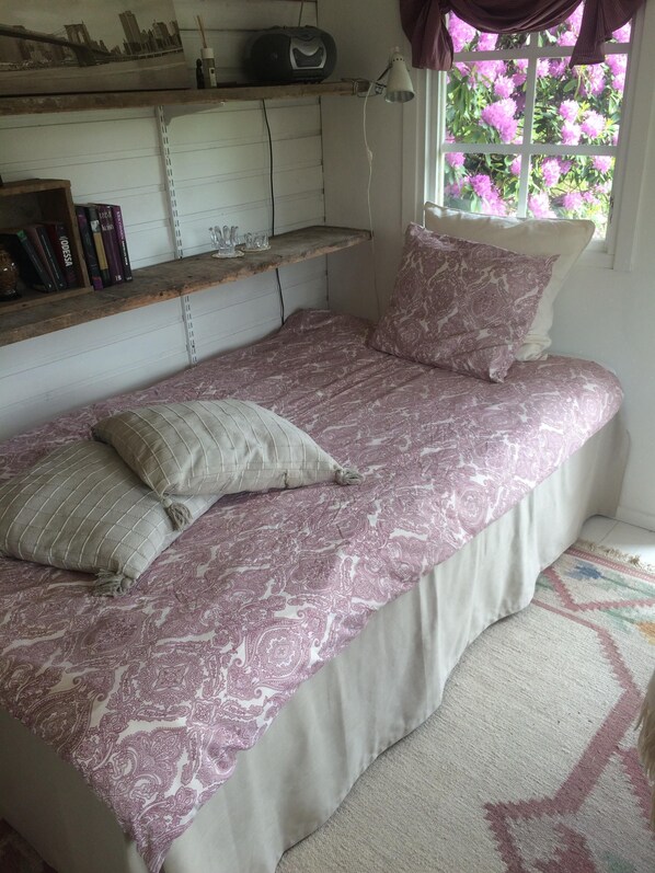 A bedroom with a bed, pillows, a shelf with books, a lamp, and a window with flowers. - Swedish Idyll (Falkenberg)