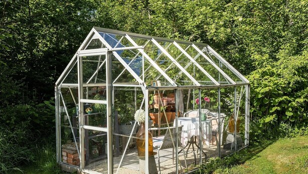 A glass greenhouse with a triangular roof, containing a table and chairs, surrounded by plants and gardening tools. - Swedish Idyll (Falkenberg)