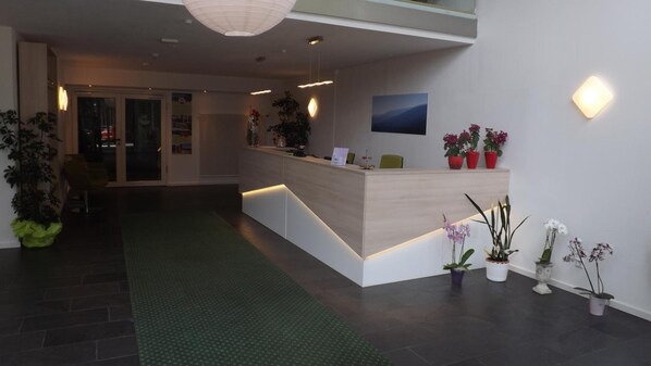 A modern reception area with a wooden reception desk, potted plants, and a green carpet.