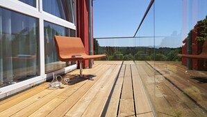 Una terraza de madera con una única silla naranja, una mesa de vidrio y una jarra de vidrio transparente.
