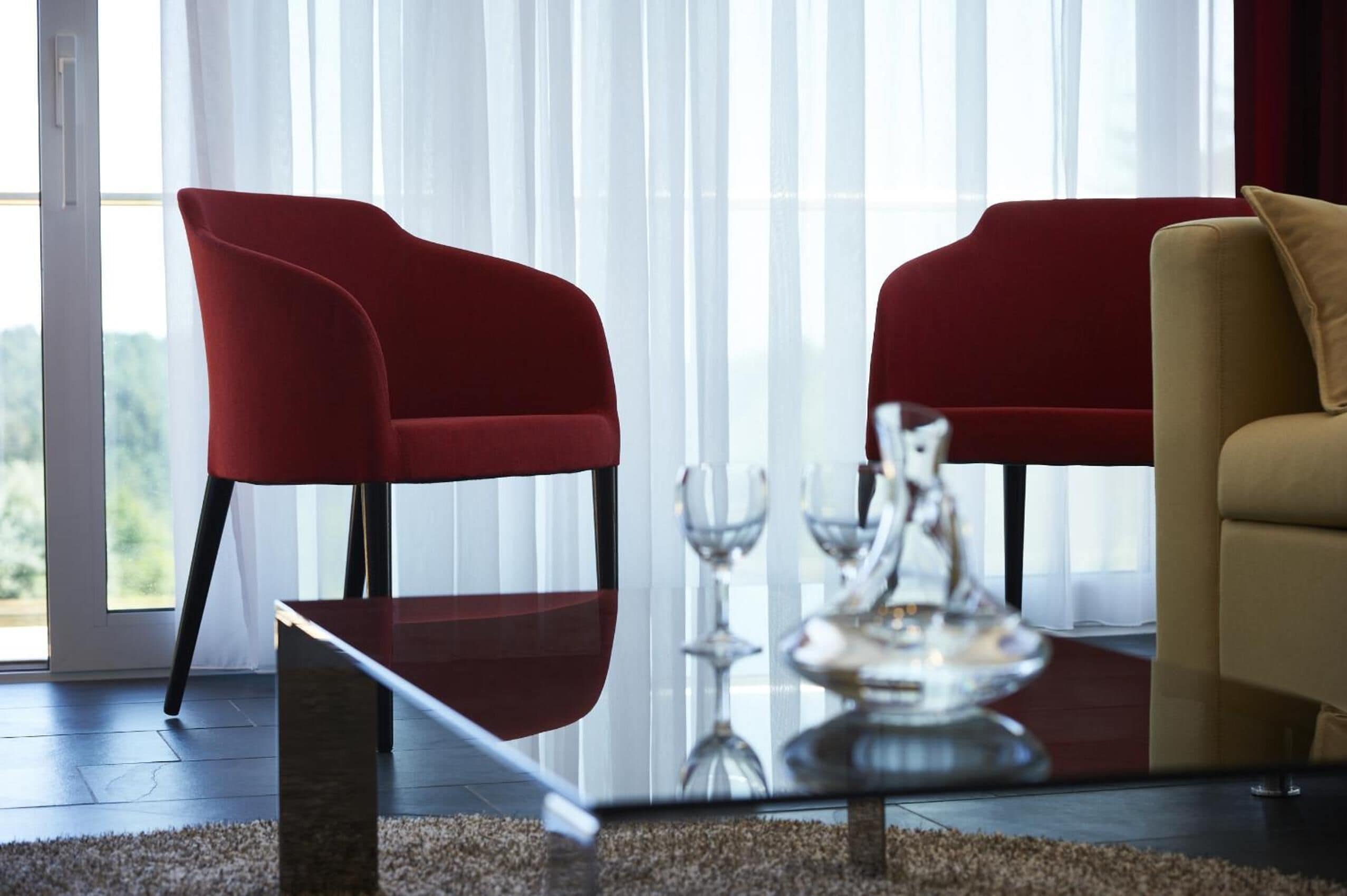 Een moderne woonkamer met twee rode fauteuils en een beige bank, een glazen salontafel, en uitzicht op groen door grote ramen.