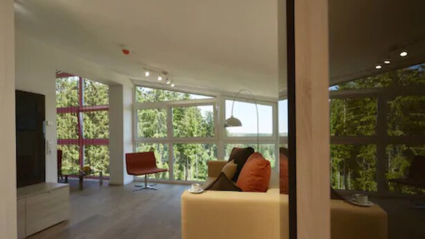 Una sala de estar moderna con un sofá, una silla y un televisor. Amplias ventanas ofrecen vistas a árboles.