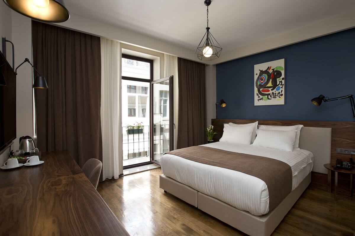 A hotel room with a large bed, a desk with a kettle, a chair, and a view through a sliding glass door.