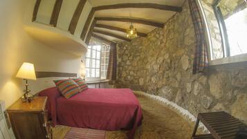 A bedroom with a stone wall, a wooden ceiling, a bed with a red bedspread, a nightstand with a lamp, and a window with curtains.