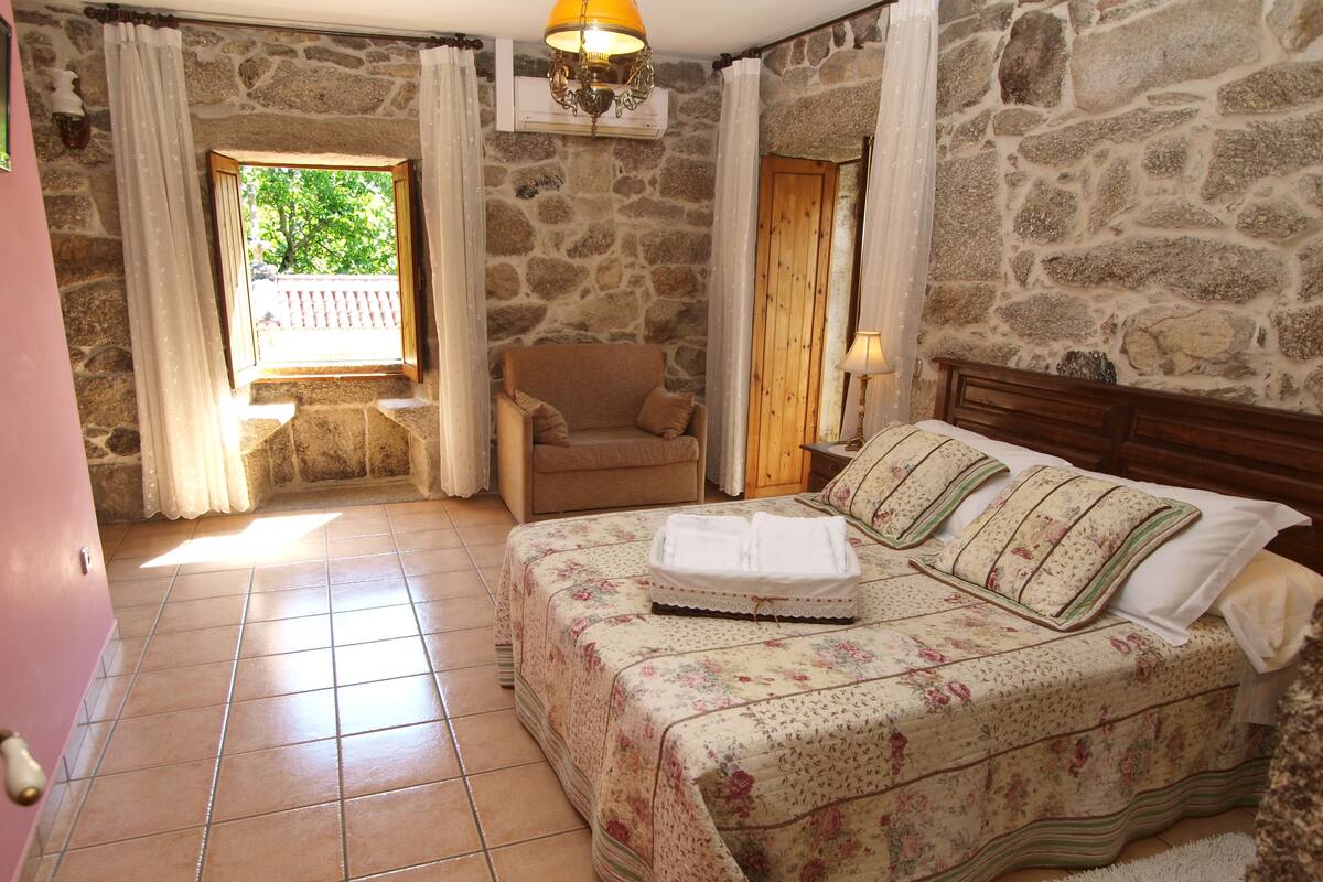 A bedroom with a stone wall, a bed with floral bedding, a wooden headboard, a nightstand, a chair, and a window with curtains.