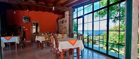 A dining area with tables set for guests, large windows offering a view of the outdoors, and a stone wall.
