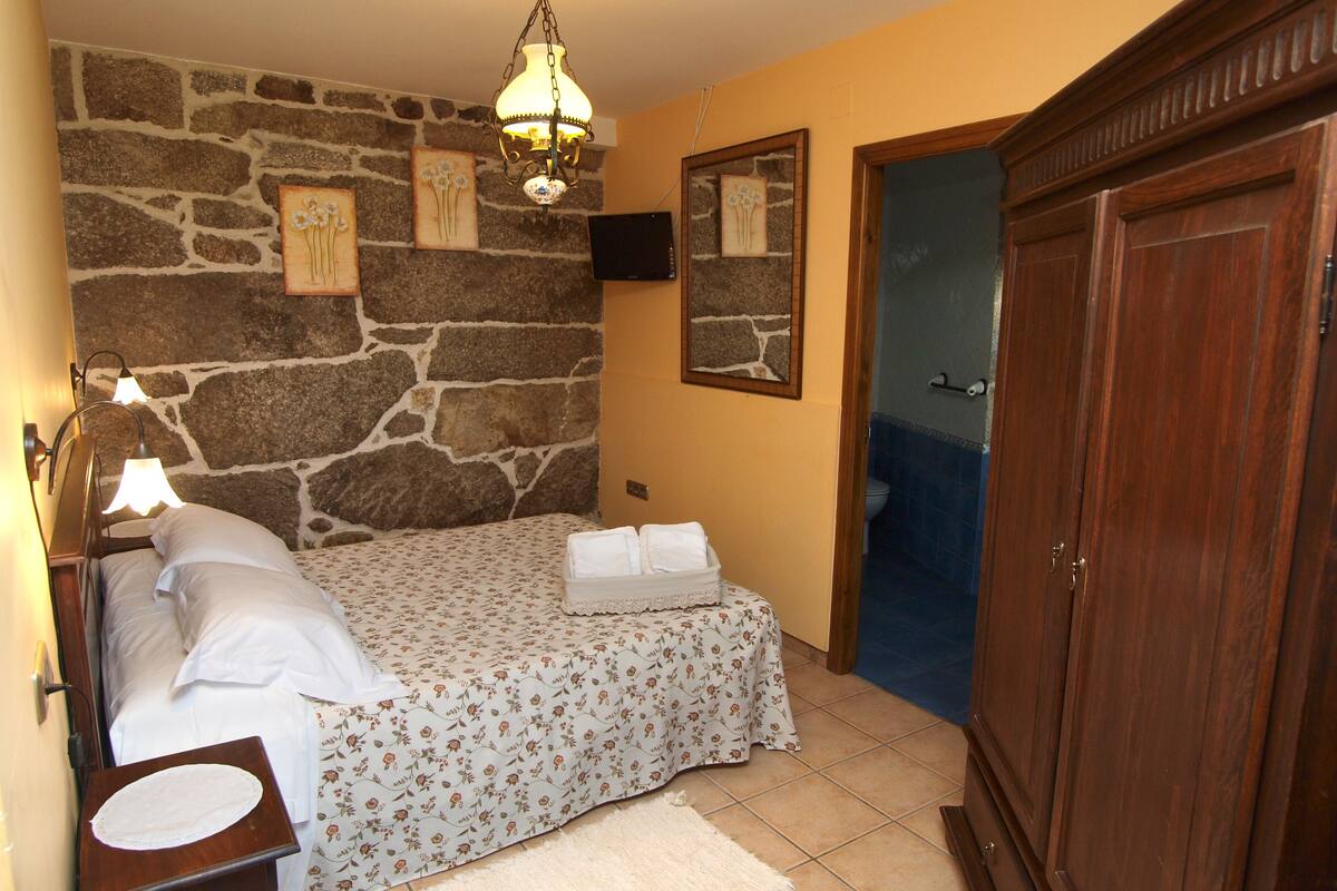 A bedroom with a stone wall, a bed with floral bedding, a bedside table with a lamp, a wooden wardrobe, and a mirror on the wall.