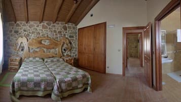 Un dormitorio con pared de piedra, techo de madera, una cama con colcha estampada, armario de madera y un baño visible a través de una puerta abierta.
