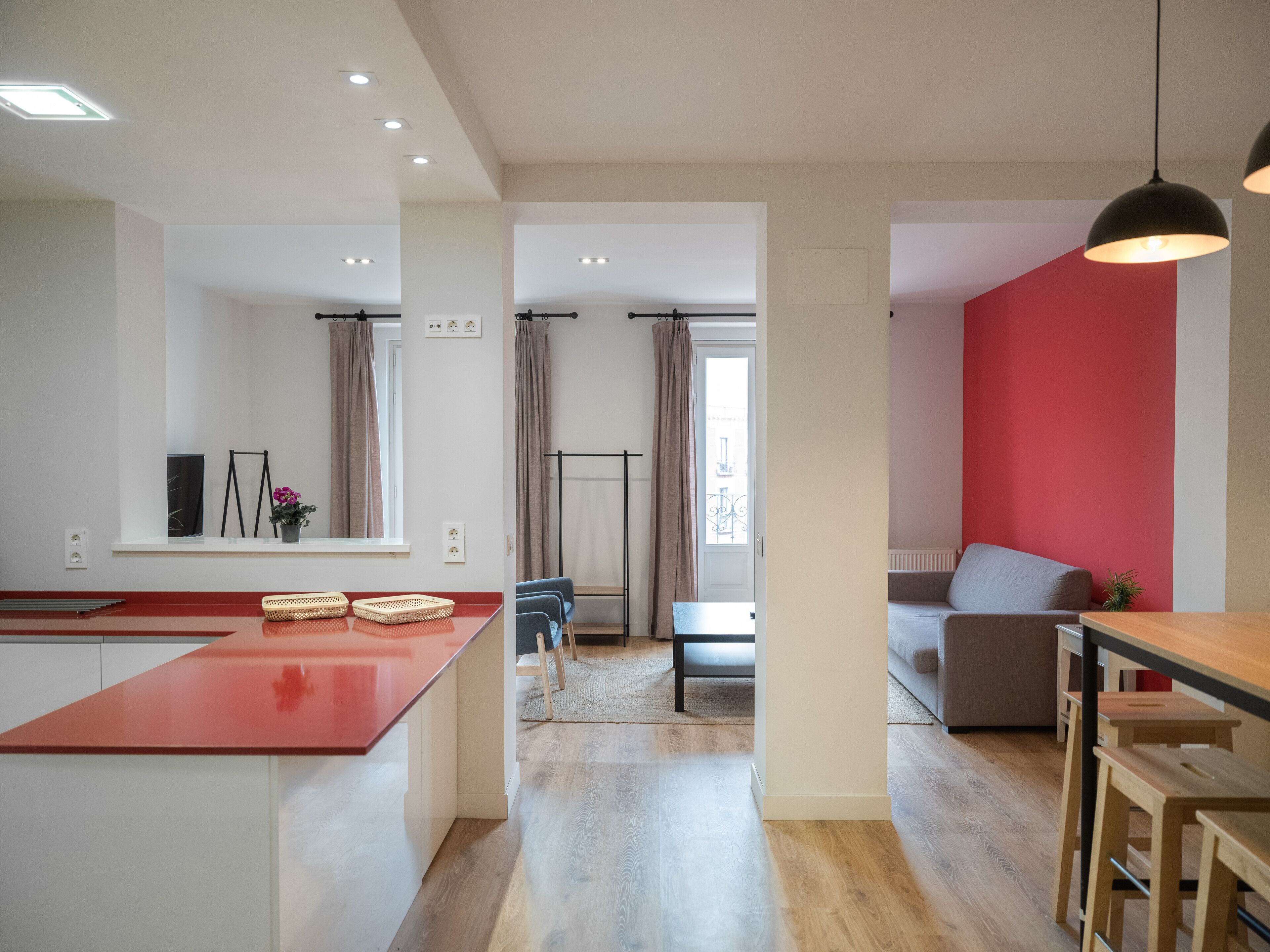 A modern kitchen with a red countertop, a dining area with a wooden table and chairs, and a living room with a red accent wall and a sofa.