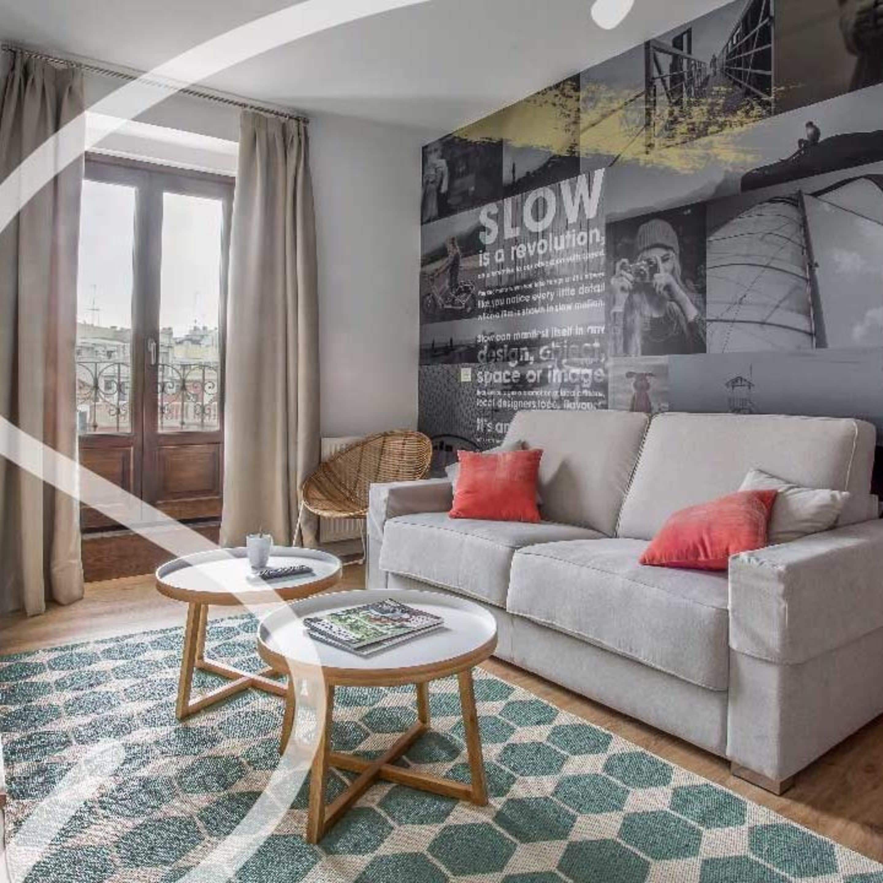 Un salon moderne avec un canapé gris, deux petites tables rondes en bois, un fauteuil en osier et une grande œuvre d’art abstraite accrochée au mur.