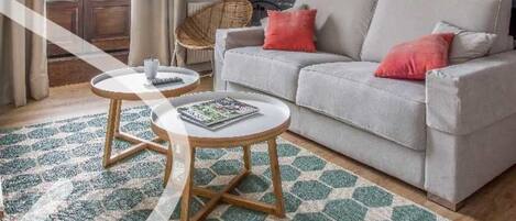 Un salon moderne avec un canapé gris, deux petites tables rondes en bois, un fauteuil en osier et une grande œuvre d’art abstraite accrochée au mur.