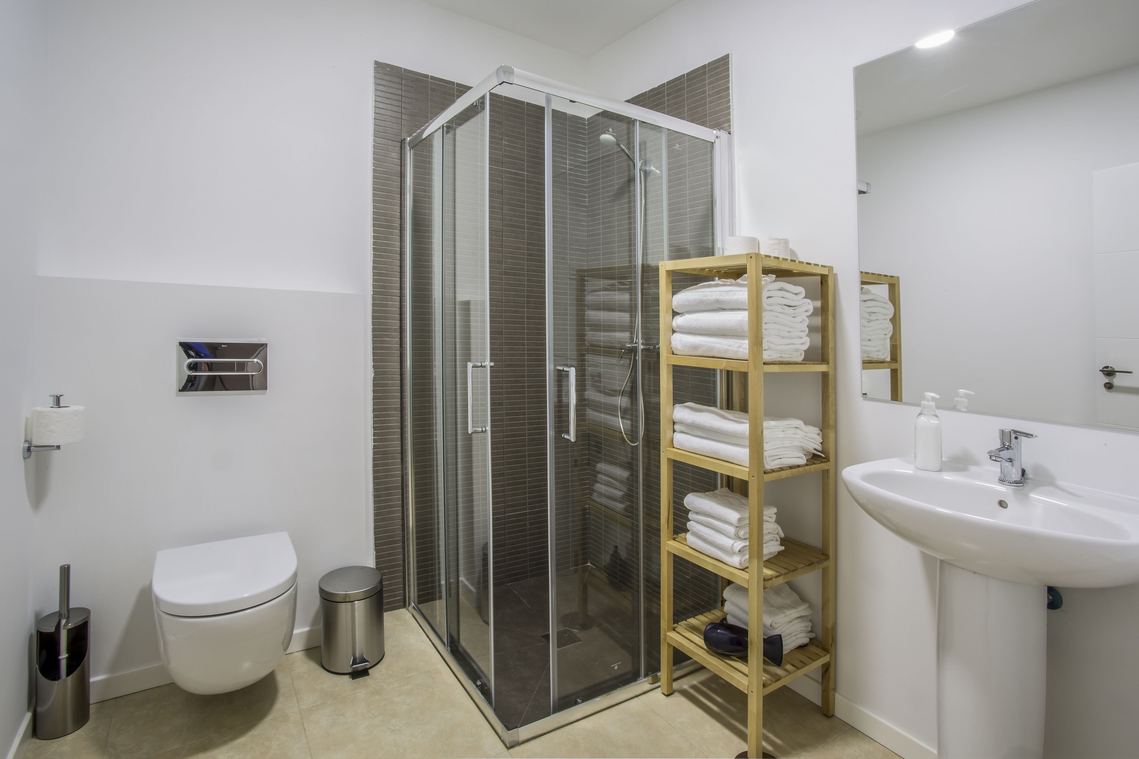 Un baño moderno con ducha de vidrio, inodoro blanco, lavamanos y estante con toallas dobladas con cuidado.