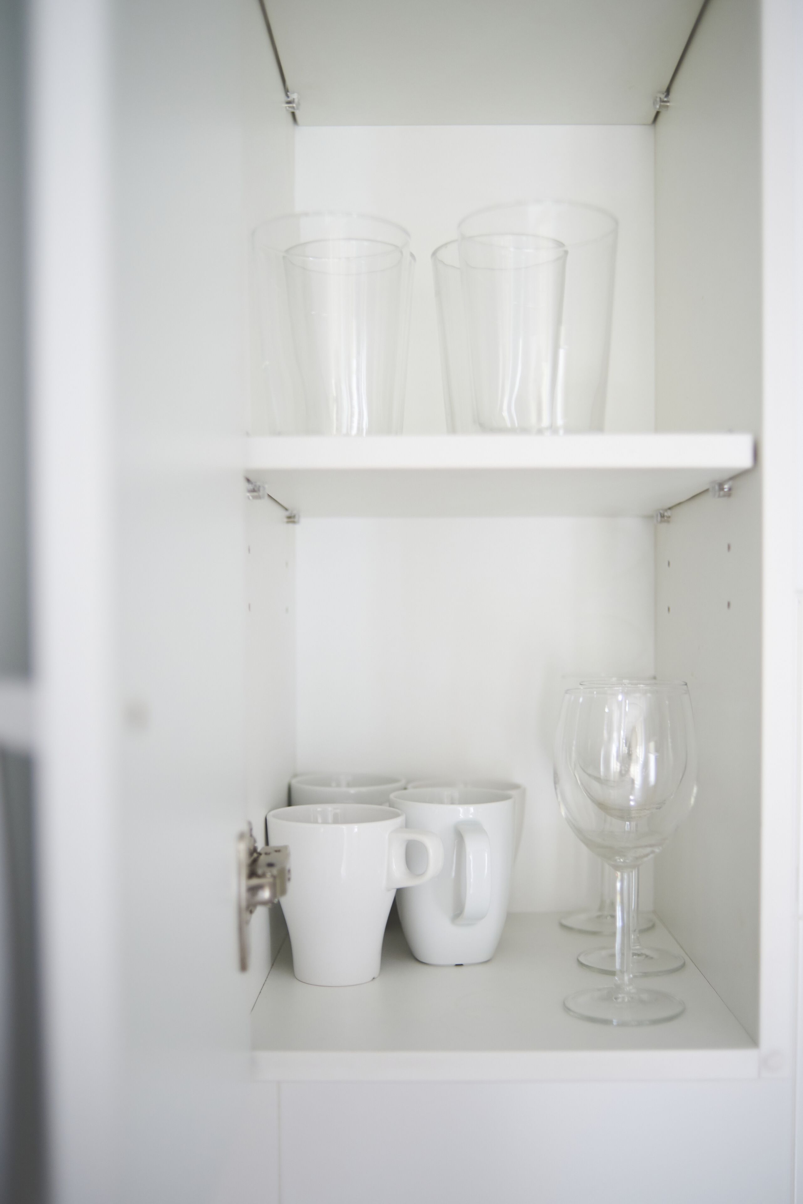 Et hvidt skab med to hylder. Den øverste hylde indeholder tre glas, og den nederste hylde har to hvide kopper og et vinglas.
