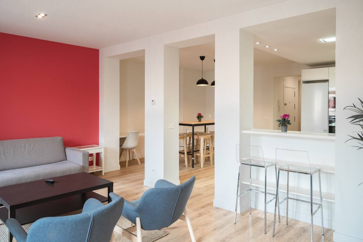 A modern living room with a grey sofa, a wooden coffee table, and blue chairs. There is a dining area with a wooden table and chairs, and a kitchen visible through an open doorway.