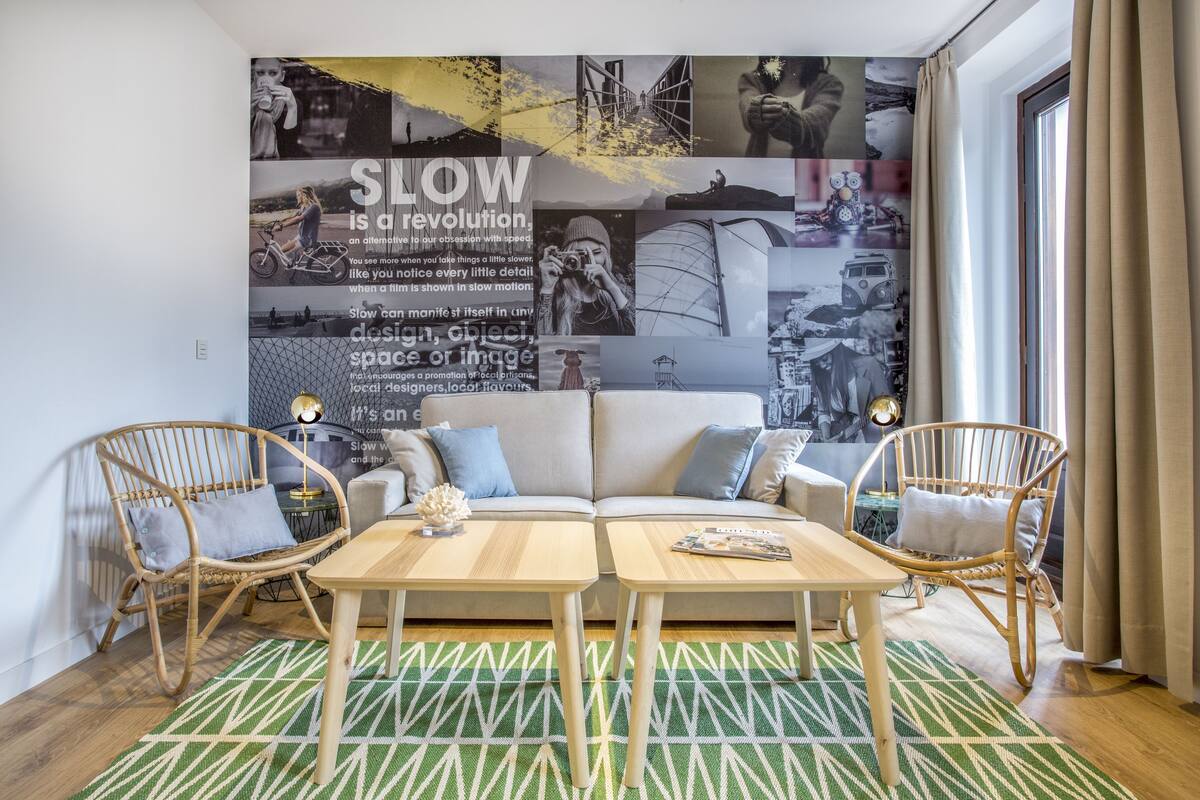 A modern living room with a large wall mural, two wooden tables, a sofa, and wicker chairs.