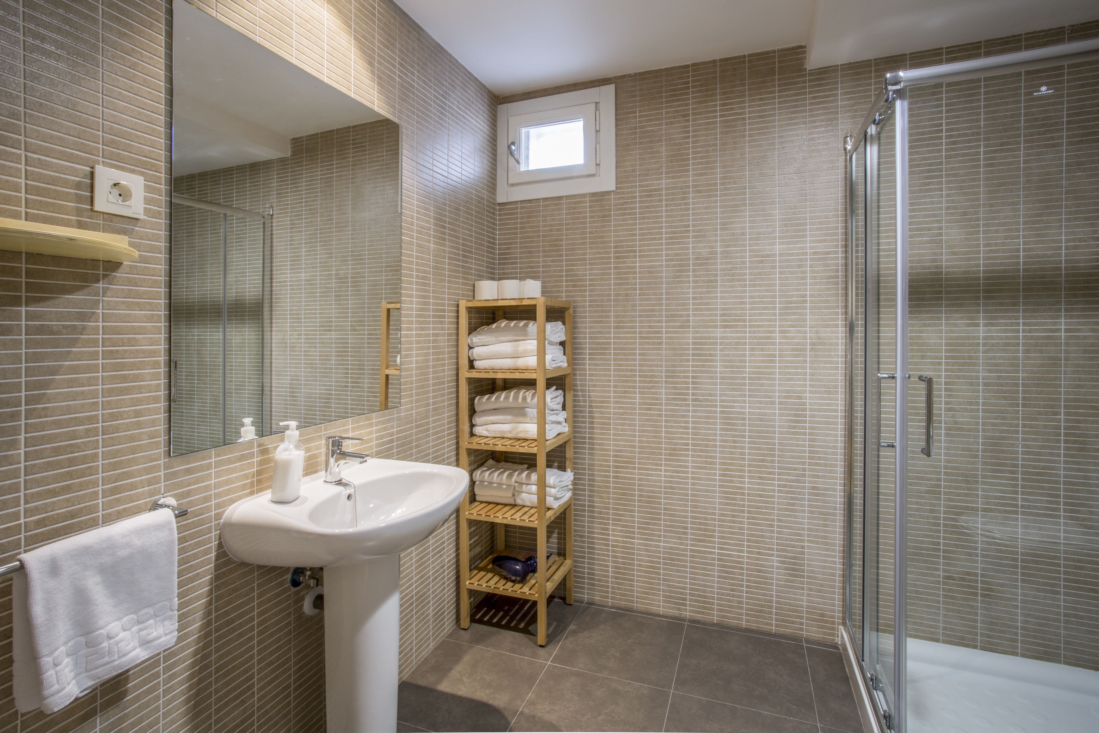 Un baño con lavamanos, espejo, toallero, estante con toallas y zona de ducha.