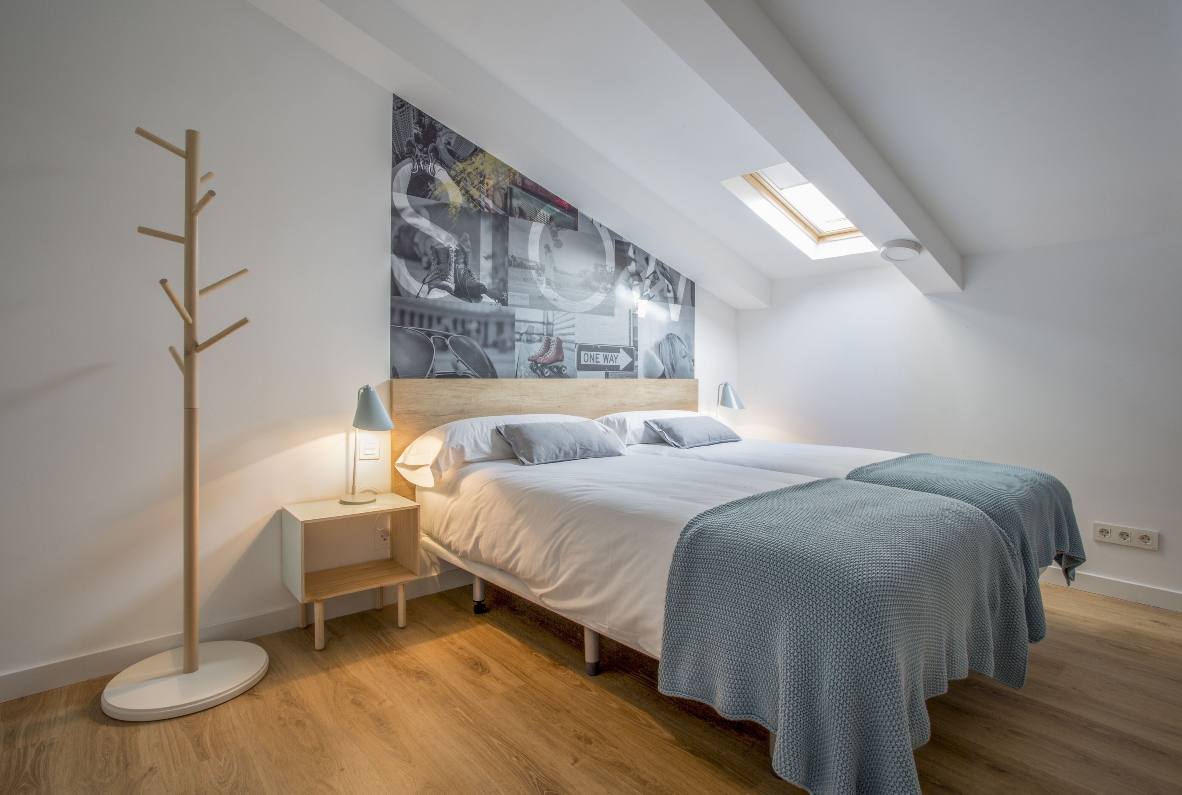 A modern bedroom with a skylight, a bed with two pillows, a bedside table with a lamp, and a wall-mounted coat rack.